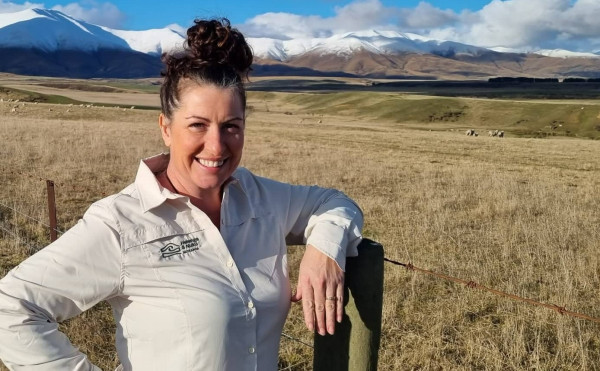 Woman wearing brnaded shirt leans on fence with mountains behind.